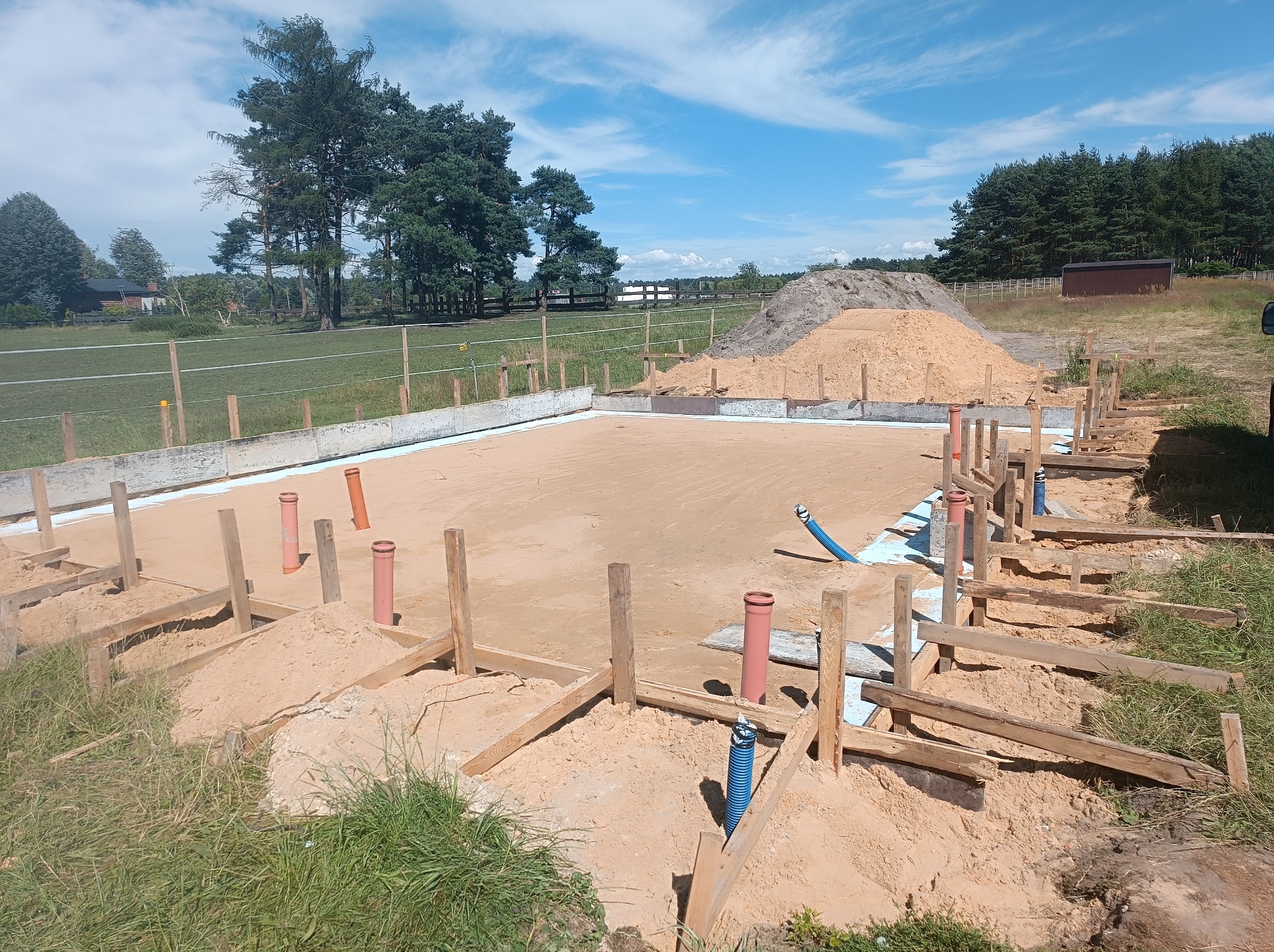 Przygotowanie terenu pod budowę: drewniane szalunki, rury instalacyjne i podsypka piaskowa na tle zieleni i błękitnego nieba.