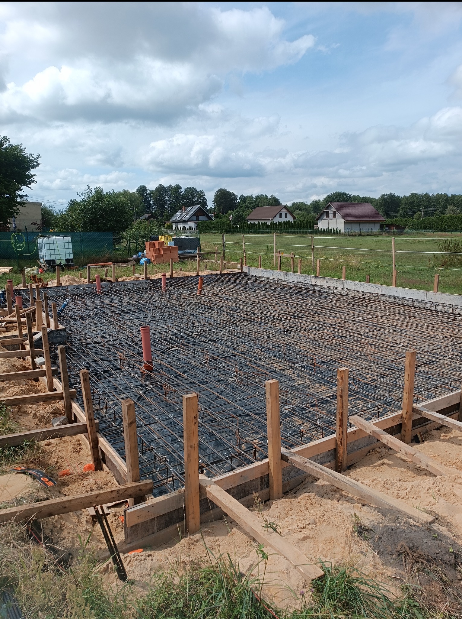 Przygotowanie zbrojenia i szalunku pod płytę fundamentową domu jednorodzinnego z widocznymi pionowymi rurami kanalizacyjnymi, w tle zabudowa jednorodzinna i błękitne niebo z chmurami.