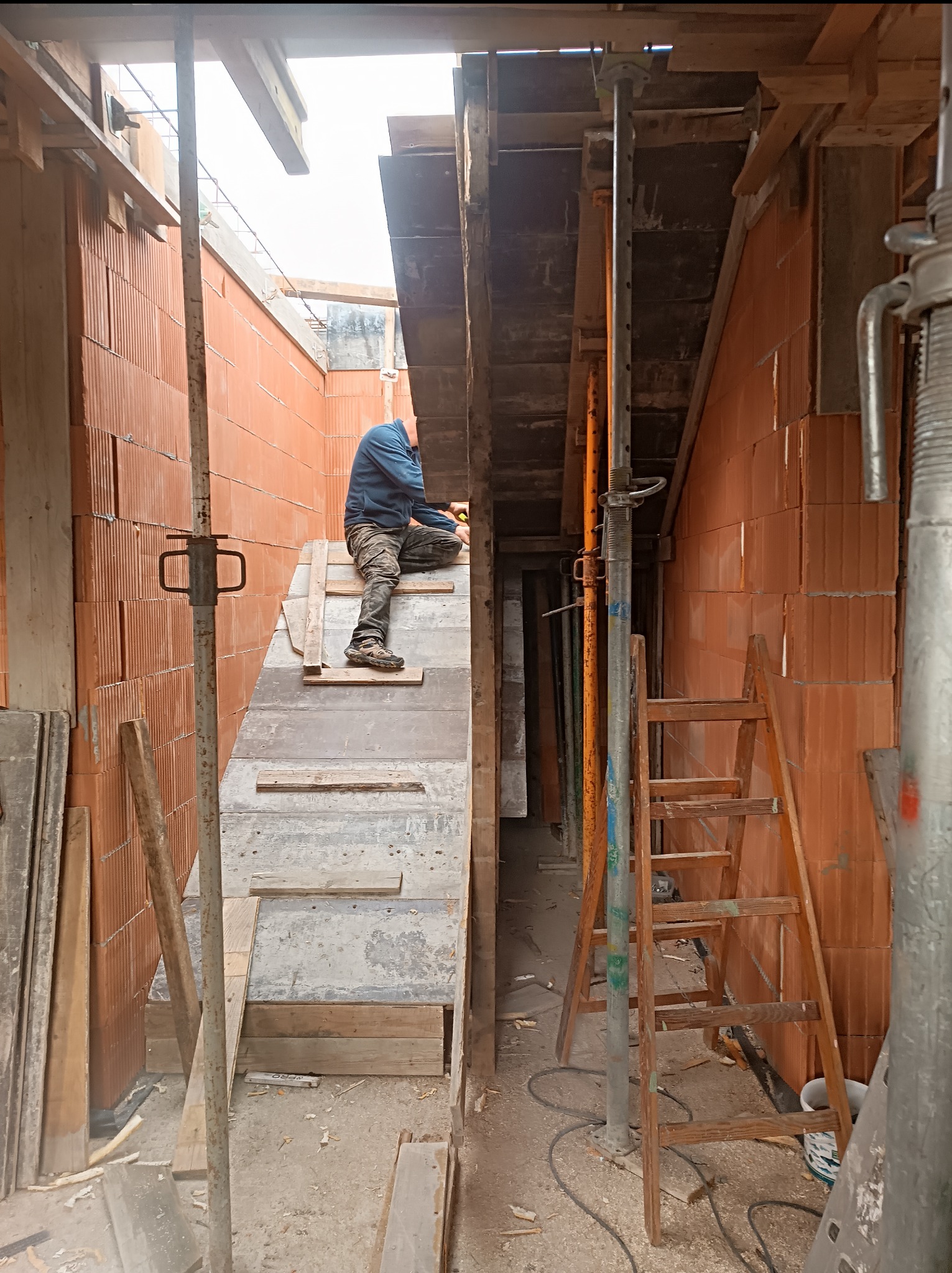 Budowa domu: murarz pracuje na drewnianym rusztowaniu wewnątrz budynku, widoczne ściany z czerwonej cegły, podpory i drewniana drabina.