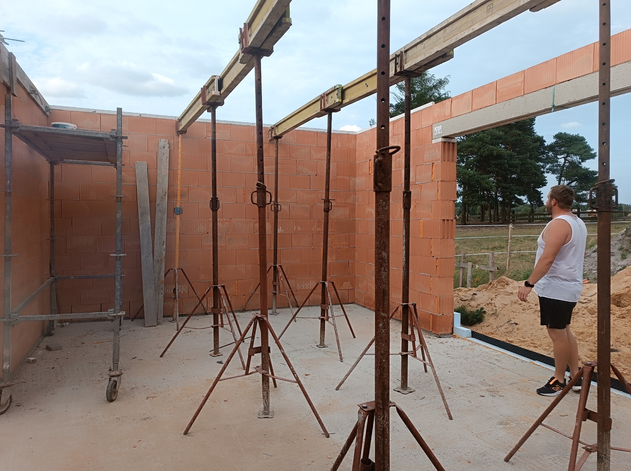 Wnętrze wznoszonego domu z czerwonej cegły ceramicznej, widoczne stemple budowlane podtrzymujące nadproża, rusztowanie i mężczyzna w tle.