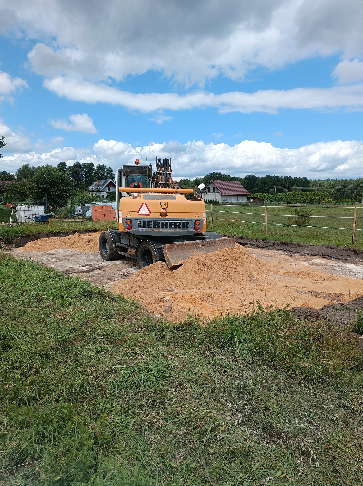 Żółta koparka Liebherr wyrównuje teren pod budowę, widoczne wykopane fundamenty i usypany piasek, w tle dom jednorodzinny i błękitne niebo z chmurami.