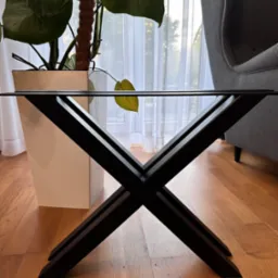 Stylowa podstawa metalowa stołu z czarnych, skrzyżowanych elementów, na drewnianej podłodze, z rośliną w tle. Minimalistyczny design.