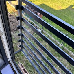 Mateusz Wadas Welding Service - Nowoczesna, grafitowa balustrada balkonowa z poziomymi szczeblami, widok z góry na trawnik i elementy ogrodu. Detal mocowania do ramy okiennej.