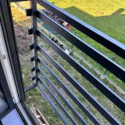 Nowoczesna, grafitowa balustrada balkonowa z poziomymi szczeblami, widok z góry na trawnik i elementy ogrodu. Detal mocowania do ramy okiennej.