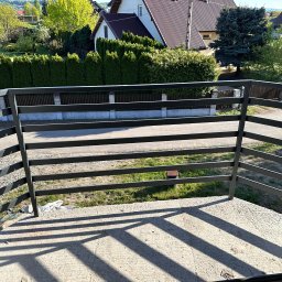 Mateusz Wadas Welding Service - Czarna balustrada balkonowa z poziomymi szczeblami, widok z góry na betonową posadzkę i zieleń. Cienie szczebli na podłodze, w tle domy i drzewa.