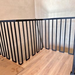 Mateusz Wadas Welding Service - Nowoczesna, czarna balustrada schodowa z pionowymi szczeblami i prostokątnymi podstawami, zamontowana w jasnym wnętrzu z drewnianą podłogą i beżowymi ścianami.