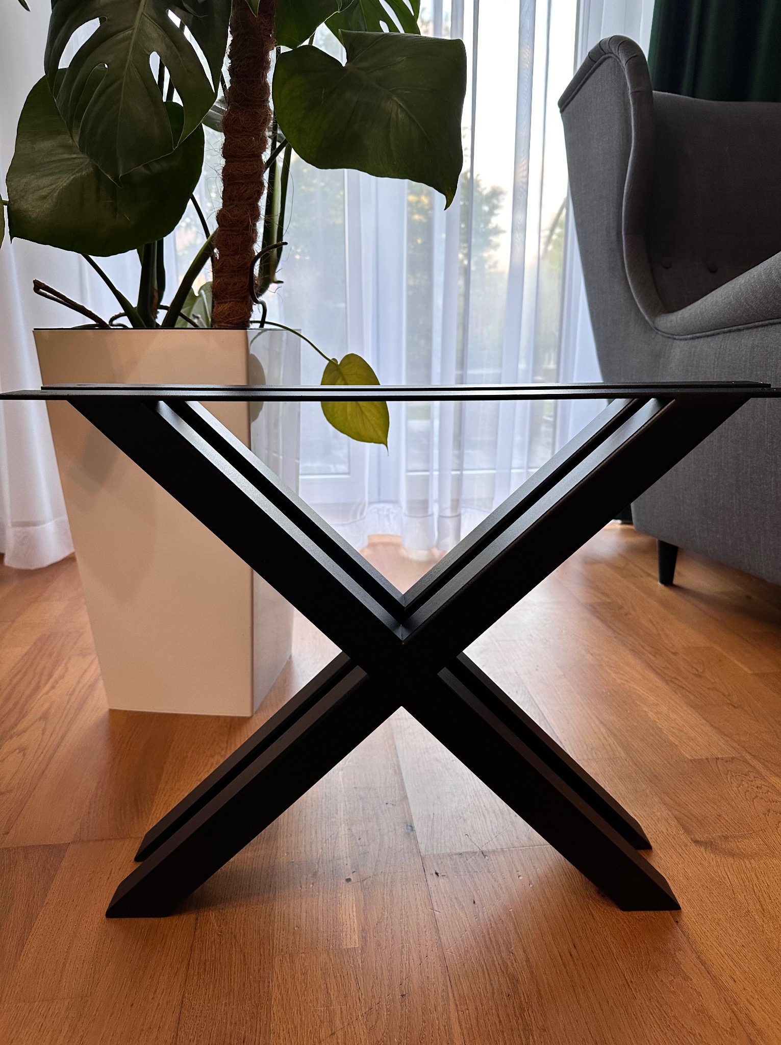 Stylowa podstawa metalowa stołu z czarnych, skrzyżowanych elementów, na drewnianej podłodze, z rośliną w tle. Minimalistyczny design.