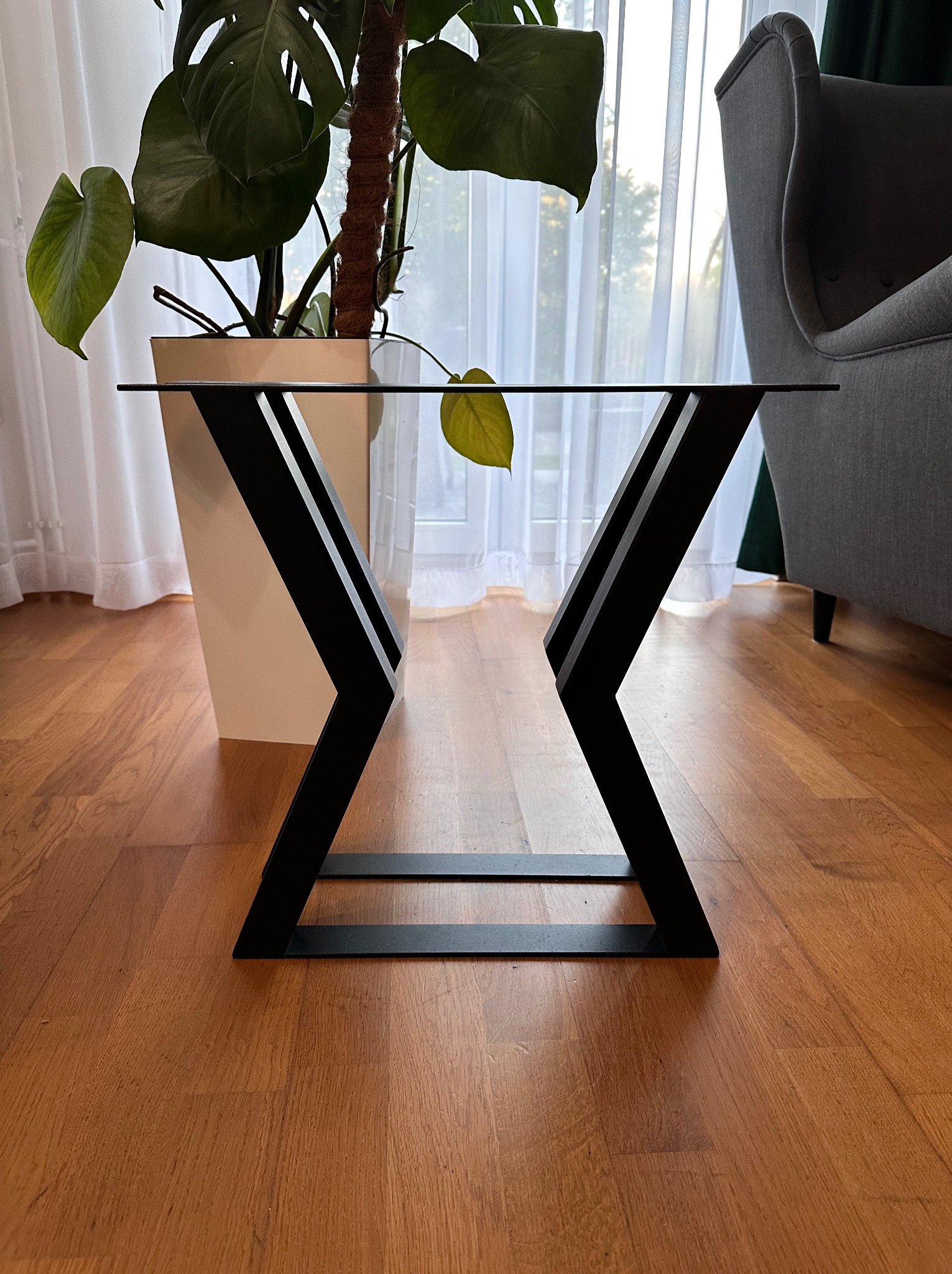 Metalowy stelaż stołu z czarnymi, geometrycznymi nogami, na drewnianej podłodze, z rośliną w tle i delikatnym światłem wpadającym przez okno.