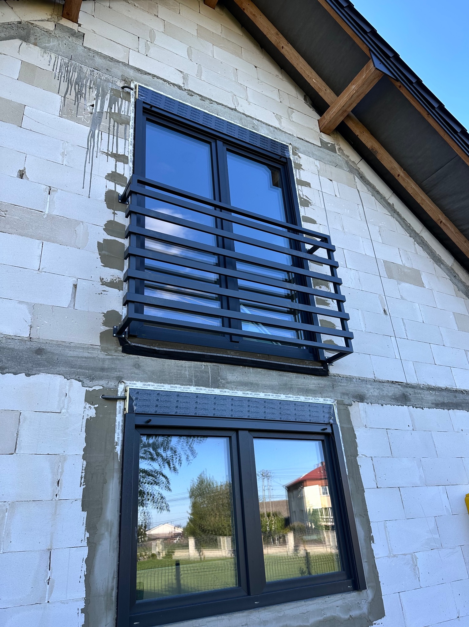 Czarna, pozioma balustrada okienna na surowej elewacji budynku z betonu komórkowego. Widoczne dwa okna z czarnymi ramami i fragment dachu.