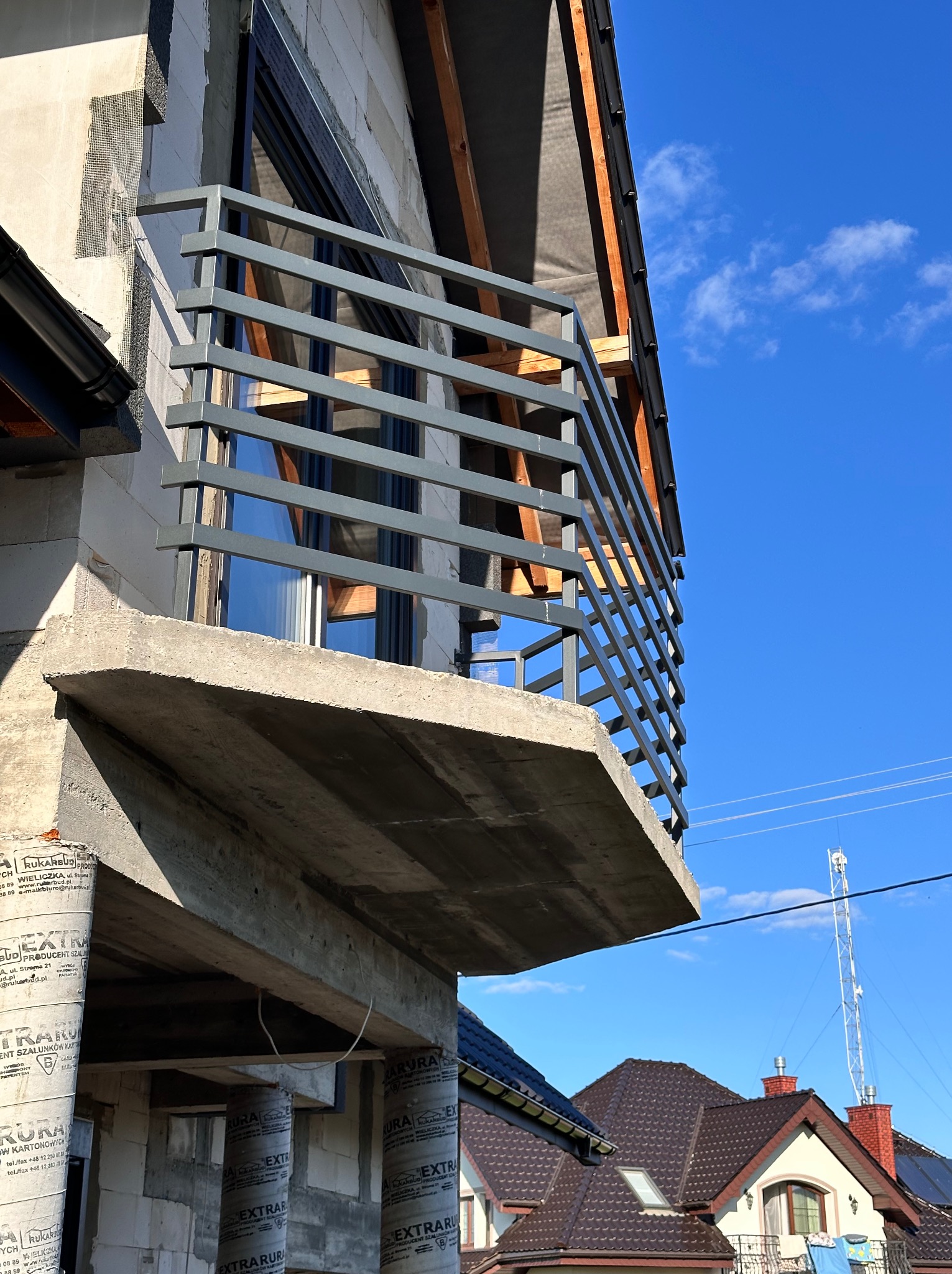 Nowoczesna balustrada balkonowa z szarego metalu na surowym balkonie w trakcie budowy, z widokiem na niebo i okoliczne dachy, w słoneczny dzień.