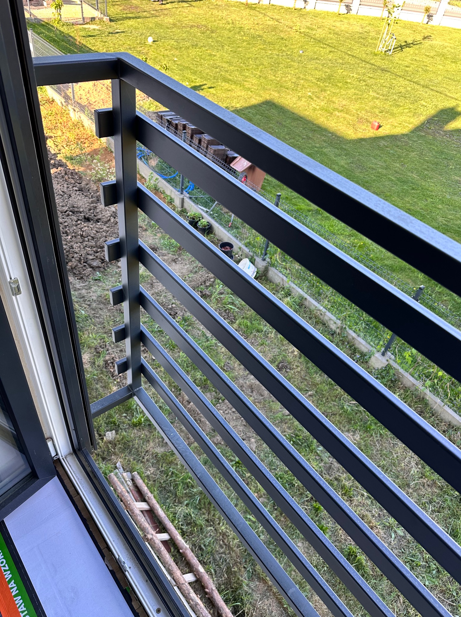 Nowoczesna, grafitowa balustrada balkonowa z poziomymi szczeblami, widok z góry na trawnik i elementy ogrodu. Detal mocowania do ramy okiennej.