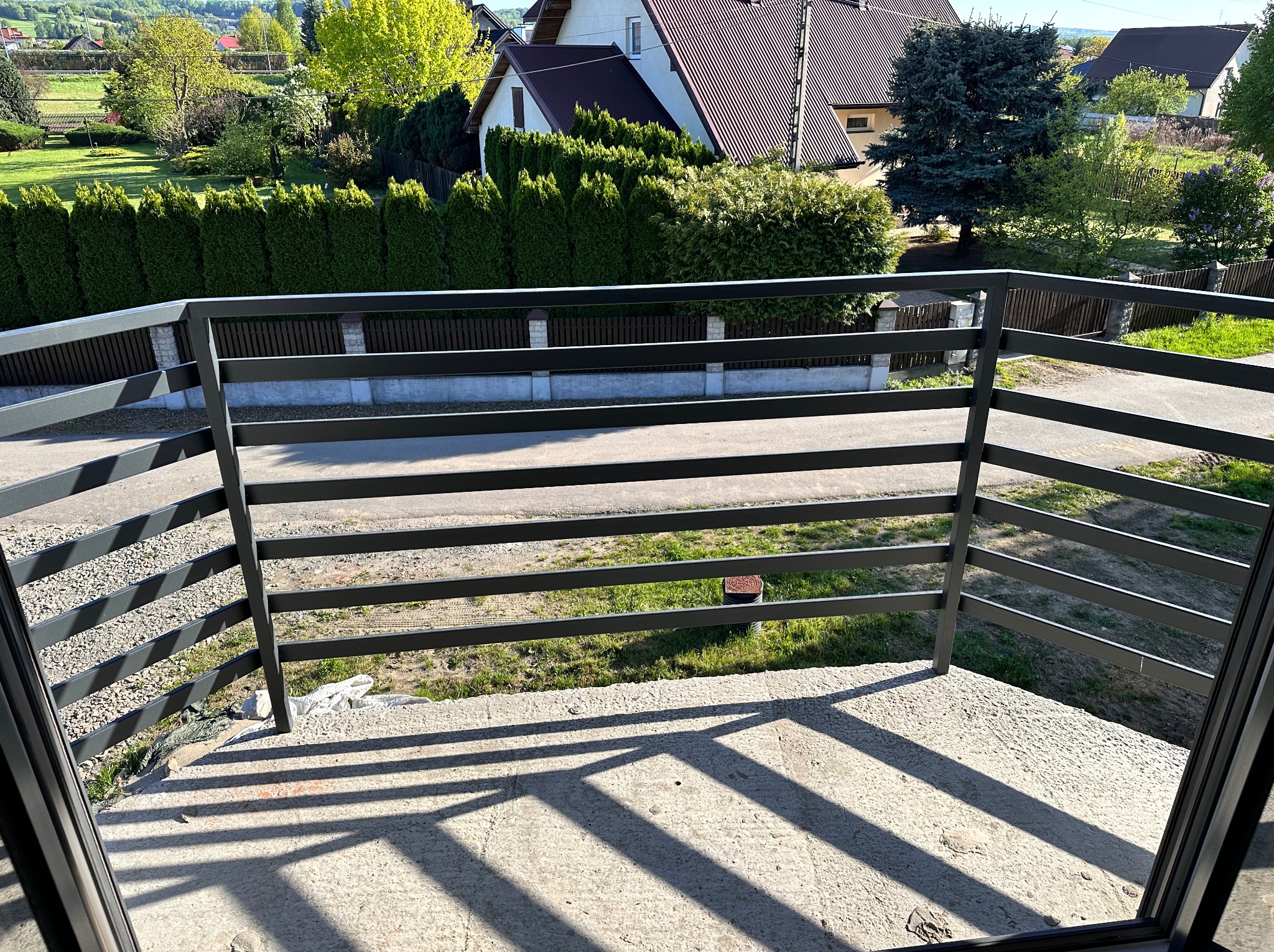 Czarna balustrada balkonowa z poziomymi szczeblami, widok z góry na betonową posadzkę i zieleń. Cienie szczebli na podłodze, w tle domy i drzewa.