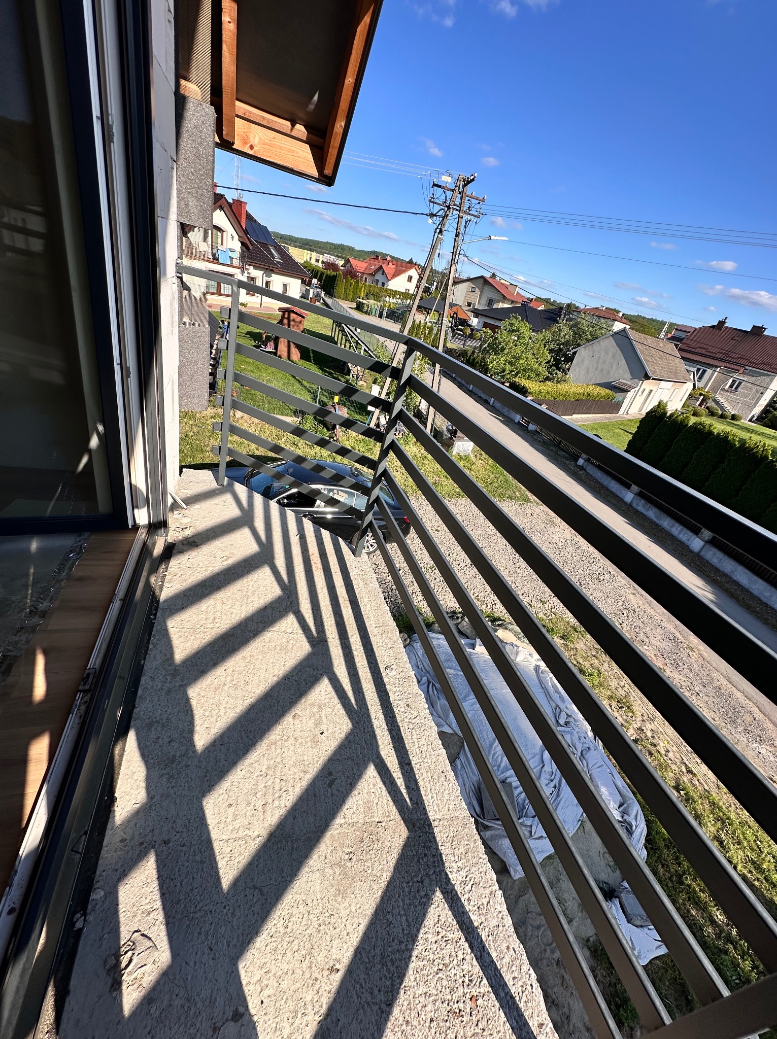 Nowoczesna balustrada balkonowa z metalu w Nagawczynie, rzucająca geometryczne cienie na betonową posadzkę. Widok na okoliczną zabudowę i błękitne niebo.