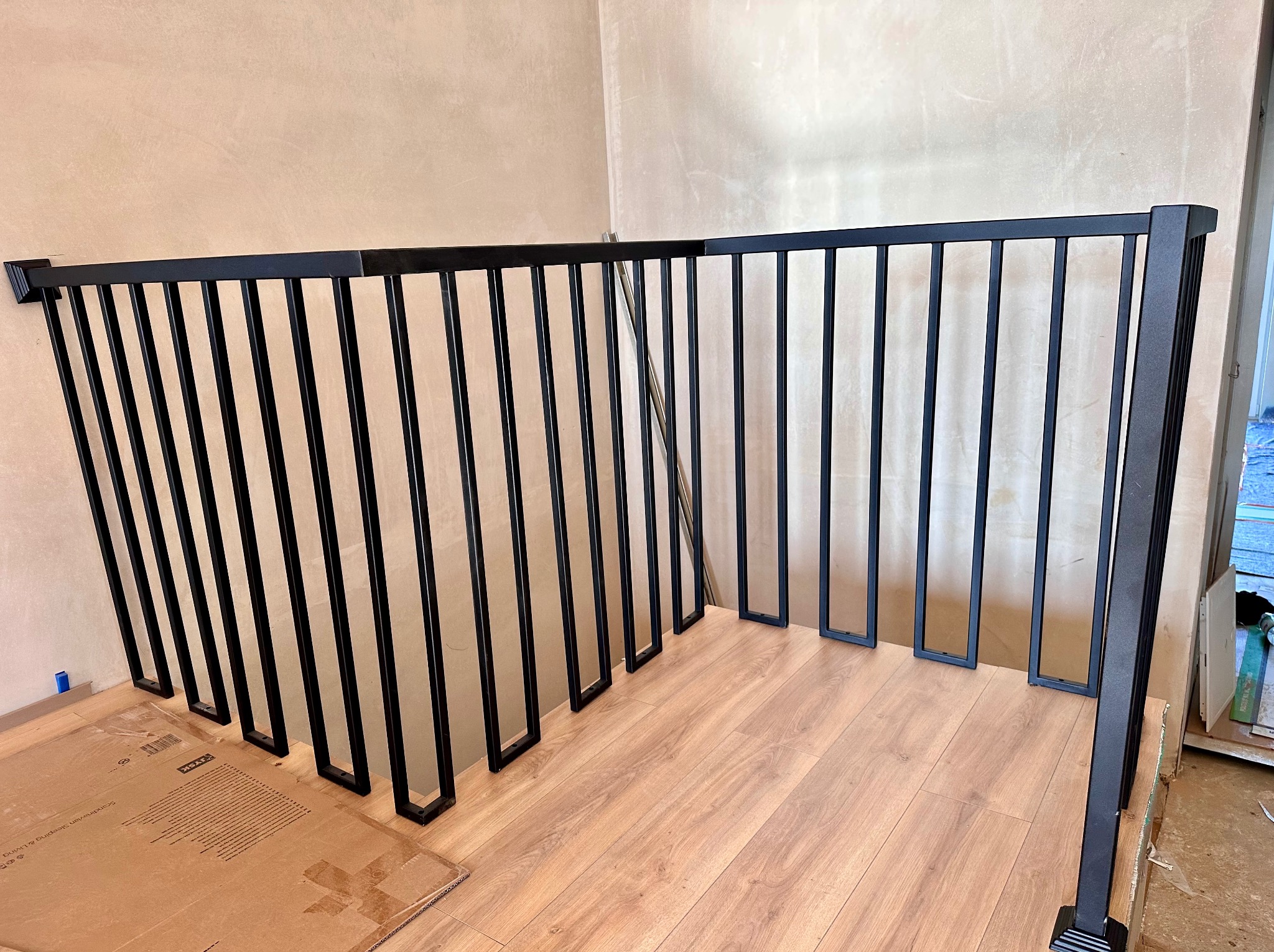 Nowoczesna, czarna balustrada schodowa z pionowymi szczeblami i prostokątnymi podstawami, zamontowana w jasnym wnętrzu z drewnianą podłogą i beżowymi ścianami.