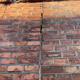 Pęknięcie pionowe biegnące przez ścianę z czerwonej cegły, widoczne uszkodzenia i nierówności muru, górą fragment instalacji elektrycznej.