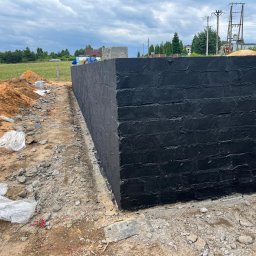 Paweł Gerega - Zabezpieczona, czarną powłoką hydroizolacyjną, ściana fundamentowa z cegły na placu budowy, widoczne narzędzia i materiały budowlane.