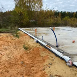Wylana płyta fundamentowa z izolacją pionową, widoczne wystające rury instalacyjne, teren wokół z piaskiem.