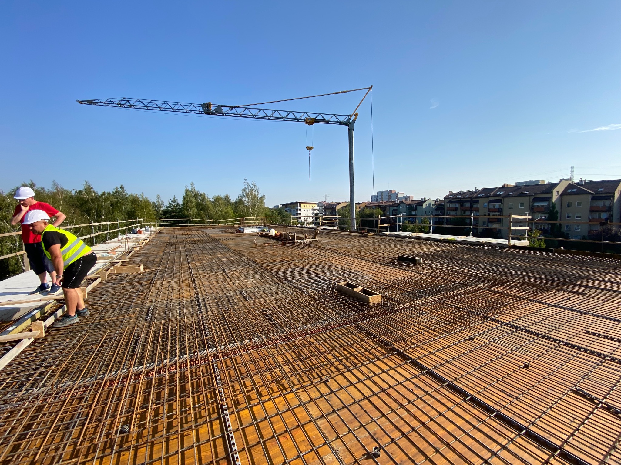 Zbrojenie stropu na budowie wielorodzinnej, widoczny żuraw budowlany i dwóch pracowników w kaskach i kamizelkach odblaskowych.