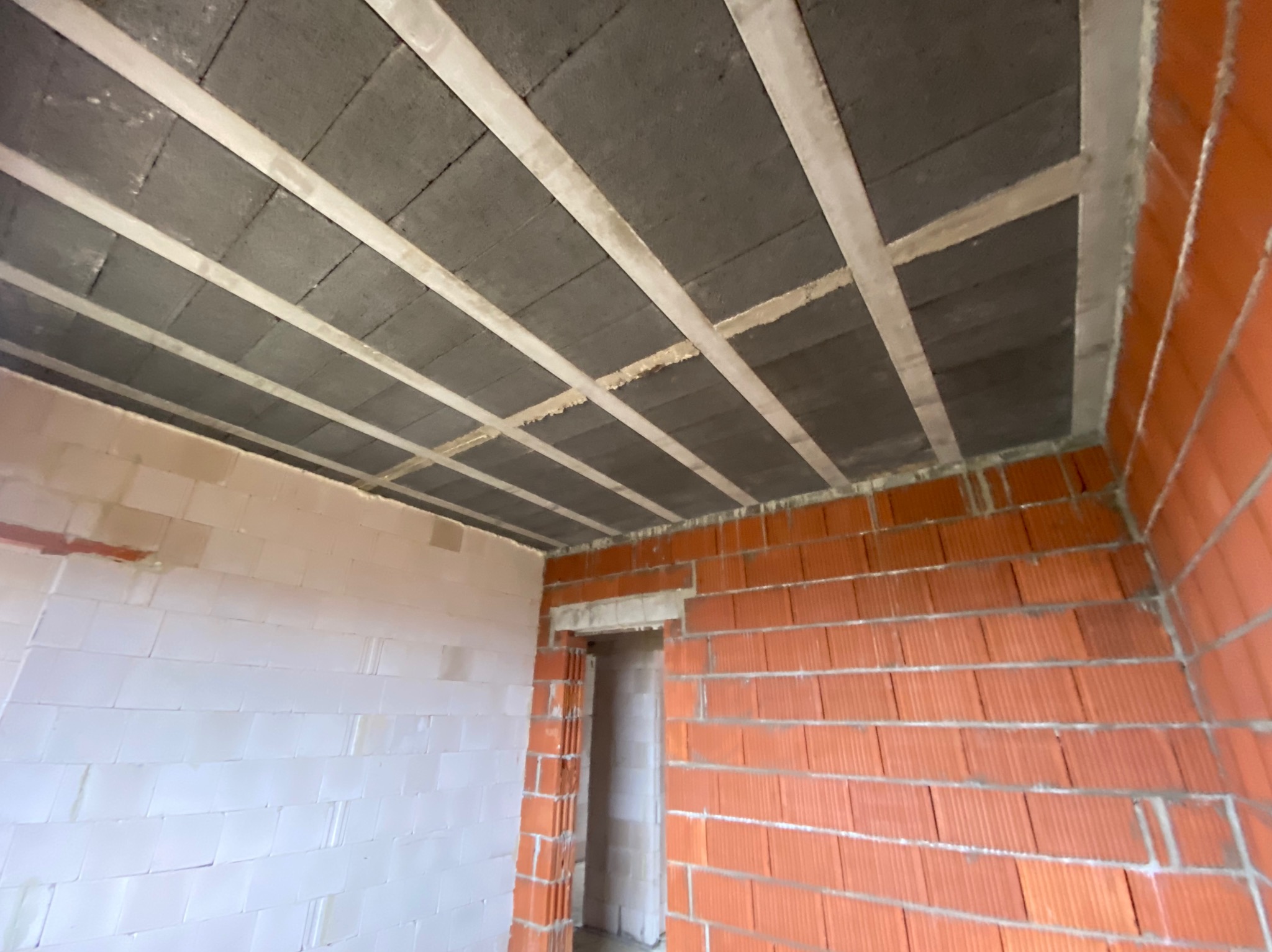 Surowy stan wnętrza budynku w trakcie budowy, widoczne ściany z czerwonej cegły dziurawki, białe ściany z bloczków oraz betonowy strop z widocznymi belkami konstrukcyjnymi.