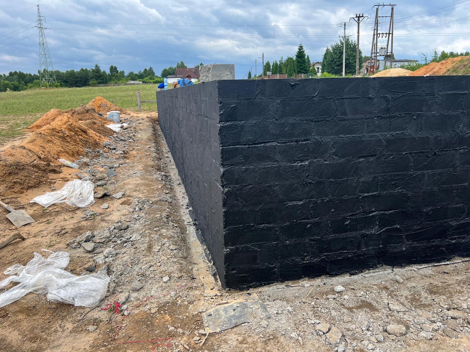 Zabezpieczona, czarną powłoką hydroizolacyjną, ściana fundamentowa z cegły na placu budowy, widoczne narzędzia i materiały budowlane.