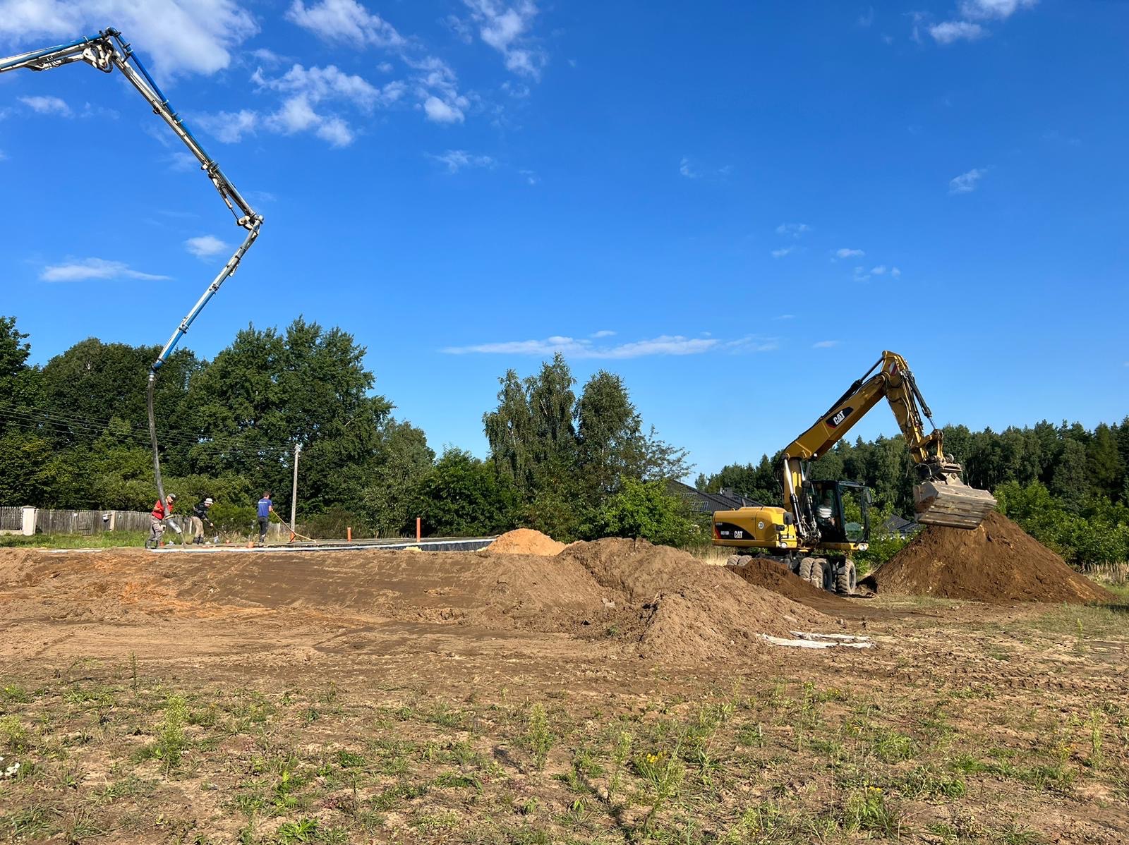 Żółta koparka CAT nabiera ziemię na placu budowy przy fundamencie, w tle widać pompę do betonu i robotników.