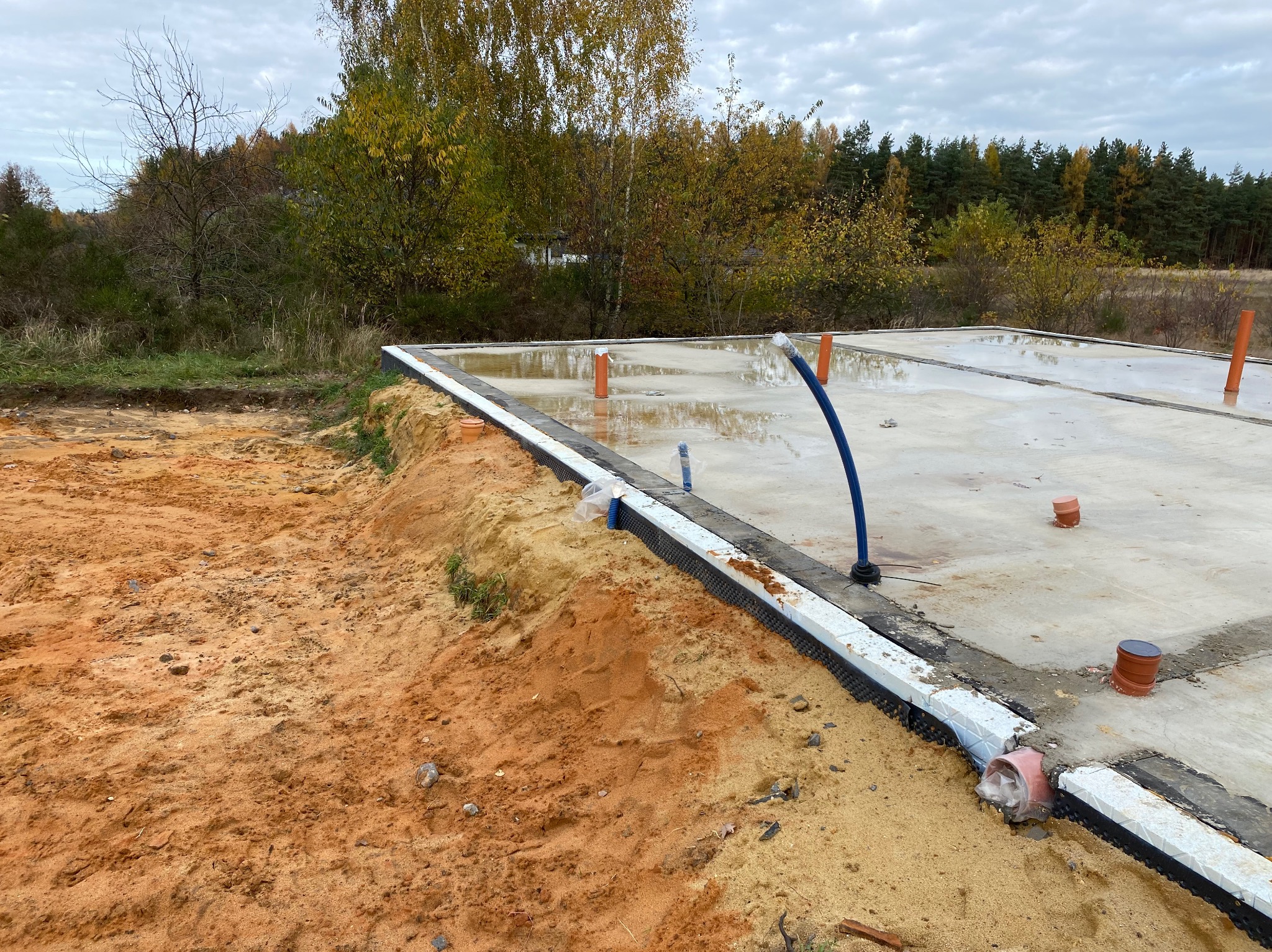 Wylana płyta fundamentowa z izolacją pionową, widoczne wystające rury instalacyjne, teren wokół z piaskiem.