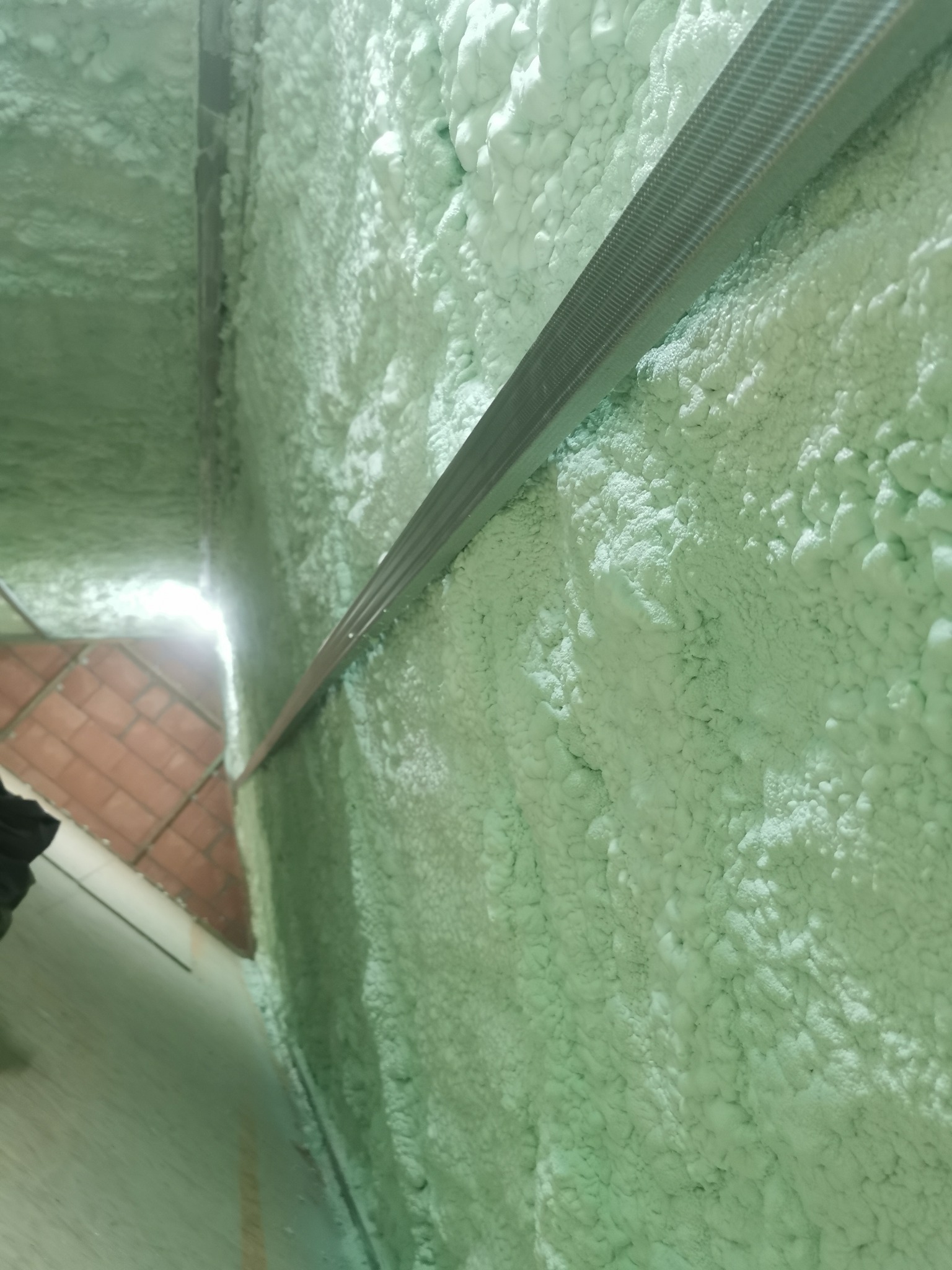 Poddasze ocieplone pianką PUR z widoczną metalową konstrukcją, jasne światło wpadające z zewnątrz. Widoczna struktura pianki i fragment podłogi z cegieł.