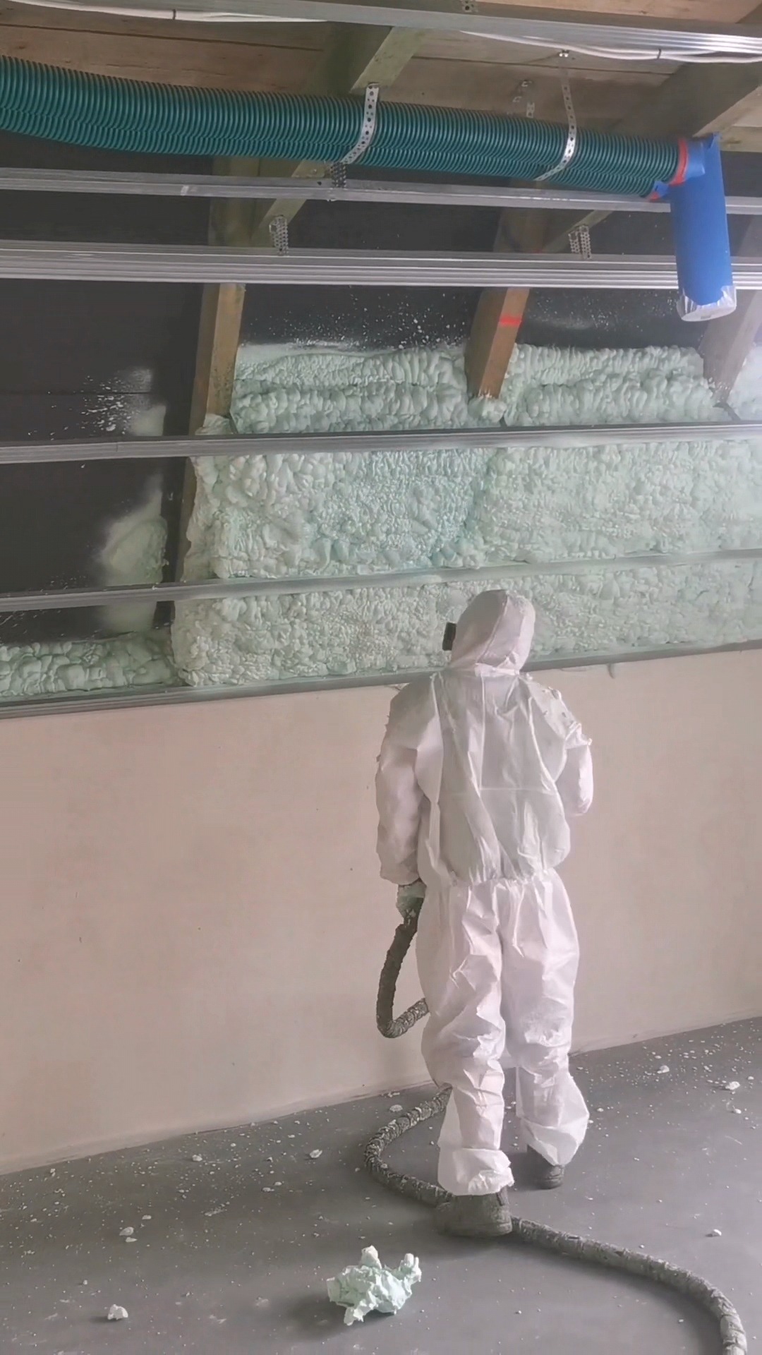 Pracownik w kombinezonie aplikuje pianę PUR na poddaszu, widoczne drewniane belki i instalacja wentylacyjna. Izolacja termiczna w trakcie realizacji.