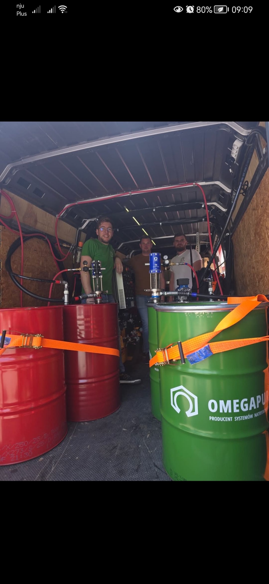 Wnętrze furgonetki z beczkami i sprzętem do natrysku pianki PUR. Widoczni trzej mężczyźni w tle, prawdopodobnie operatorzy sprzętu.