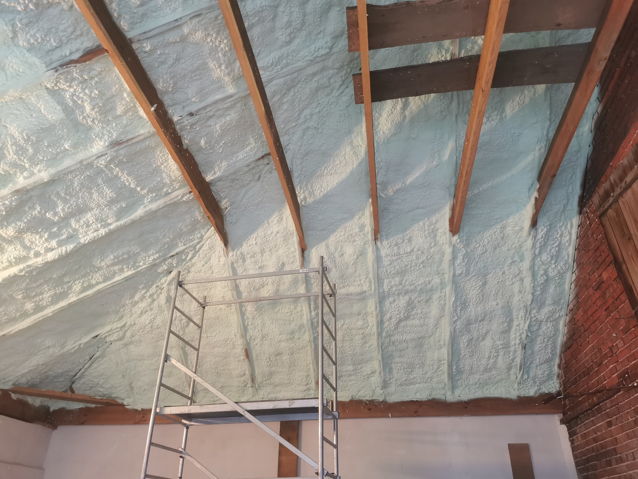 Poddasze z widoczną, świeżo nałożoną pianką izolacyjną PUR, drewnianymi belkami i fragmentem rusztowania. Ściana z czerwonej cegły w tle.