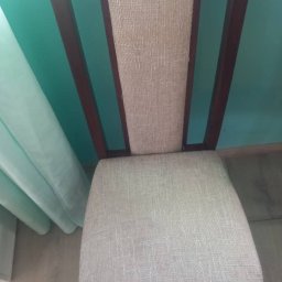 Już w porządku - Krzesło z tapicerowanym siedziskiem i oparciem, brązowe drewniane nogi, na tle turkusowej ściany i zasłony. Widok z góry na czyste krzesło.