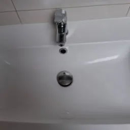 Biała umywalka z chromowaną baterią i korkiem, po sprzątaniu. Widoczne czyste płytki na ścianie. Minimalistyczny design.