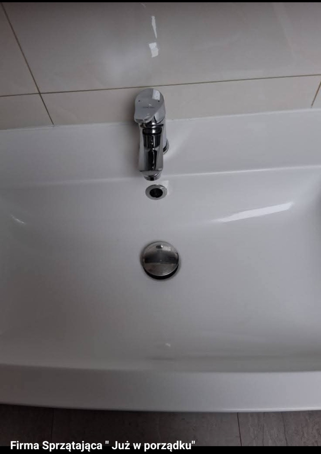 Biała umywalka z chromowaną baterią i korkiem, po sprzątaniu. Widoczne czyste płytki na ścianie. Minimalistyczny design.