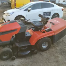 Pomarańczowo-czarny traktorek do koszenia trawy z czarnym siedzeniem, stojący na ziemi, w tle biały samochód z reklamą myjni ręcznej.