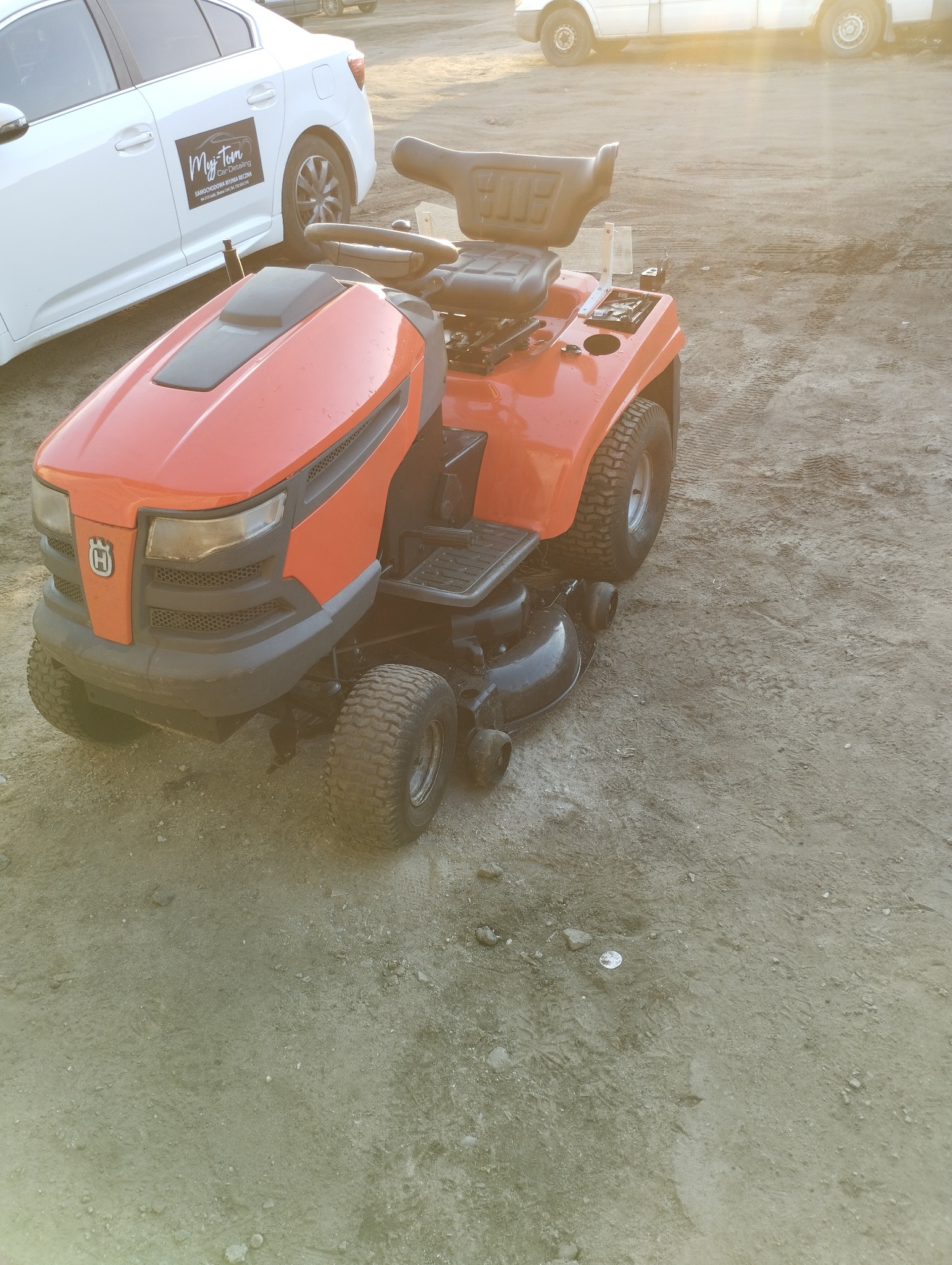 Pomarańczowy traktorek do trawy Husqvarna z czarnym siedzeniem i kierownicą, stojący na ziemi obok białego samochodu z naklejką firmy detailingowej.