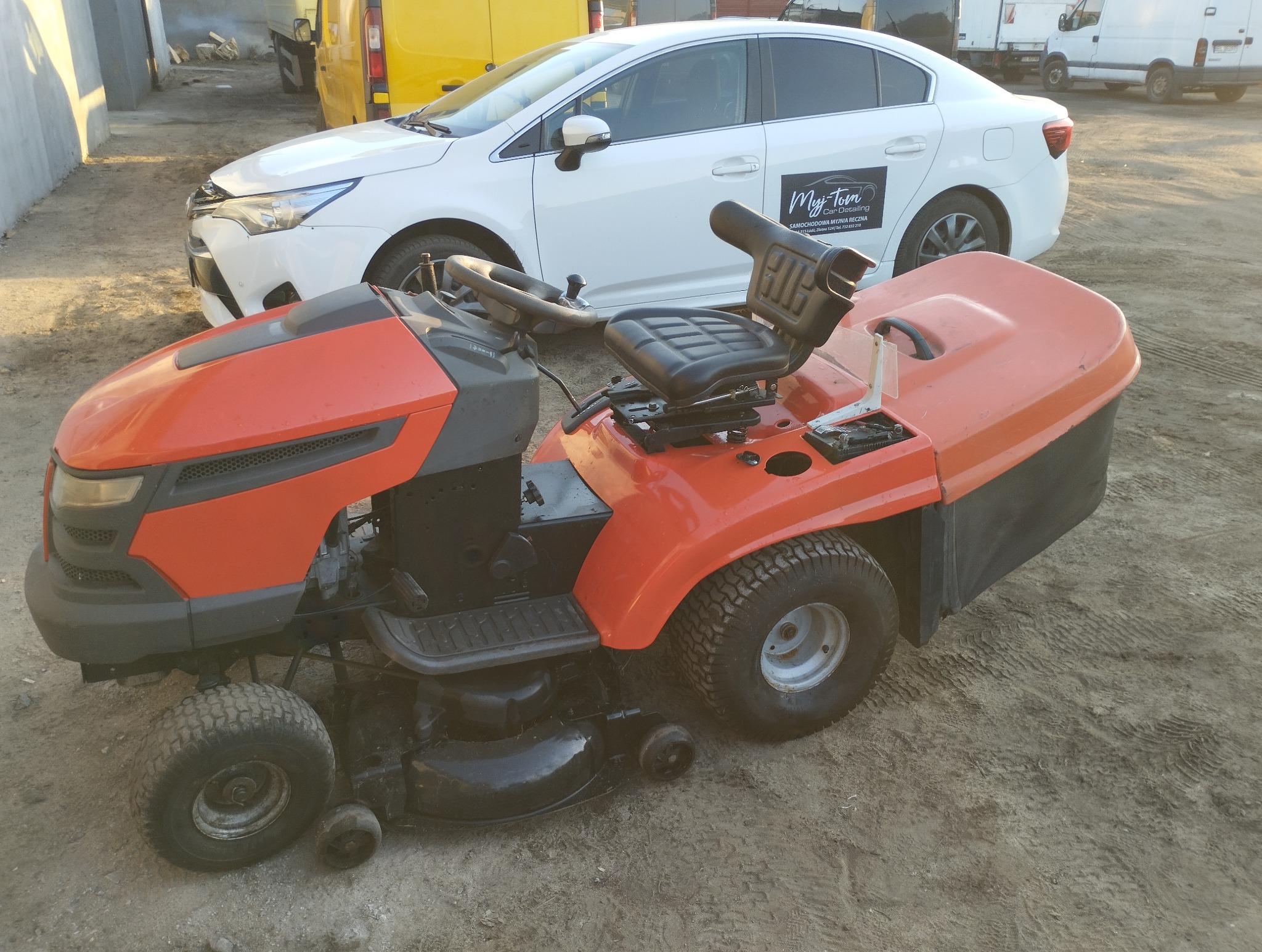 Pomarańczowo-czarny traktorek do koszenia trawy z czarnym siedzeniem, stojący na ziemi, w tle biały samochód z reklamą myjni ręcznej.