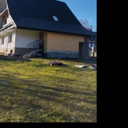 Usługi Ciesielsko-Dekarskie Łukasz Dziadoń - Dom z ciemnym, nowym dachem i oknem dachowym, widoczny z trawnika w słoneczny dzień. Część elewacji w trakcie wykończenia, widoczne palety i materiały budowlane.