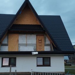Usługi Ciesielsko-Dekarskie Łukasz Dziadoń - Dom w trakcie prac dekarskich: czarna dachówka kontrastuje z jasną elewacją. Widoczne elementy konstrukcyjne dachu i rusztowanie. Tło stanowią zalesione wzgórza.