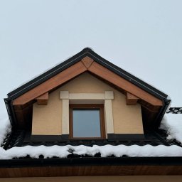 Usługi Ciesielsko-Dekarskie Łukasz Dziadoń - Zimowy widok na lukarnę z czarną dachówką i drewnianymi elementami wykończeniowymi. Śnieg na dachu i rynnach. Jasna elewacja budynku.