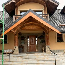 Usługi Ciesielsko-Dekarskie Łukasz Dziadoń - Dom z ciemną dachówką i drewnianym podcieniem nad wejściem, widoczne rusztowanie. Detale wykończenia z kamienia i drewna. Schody wejściowe z jasnych płytek.