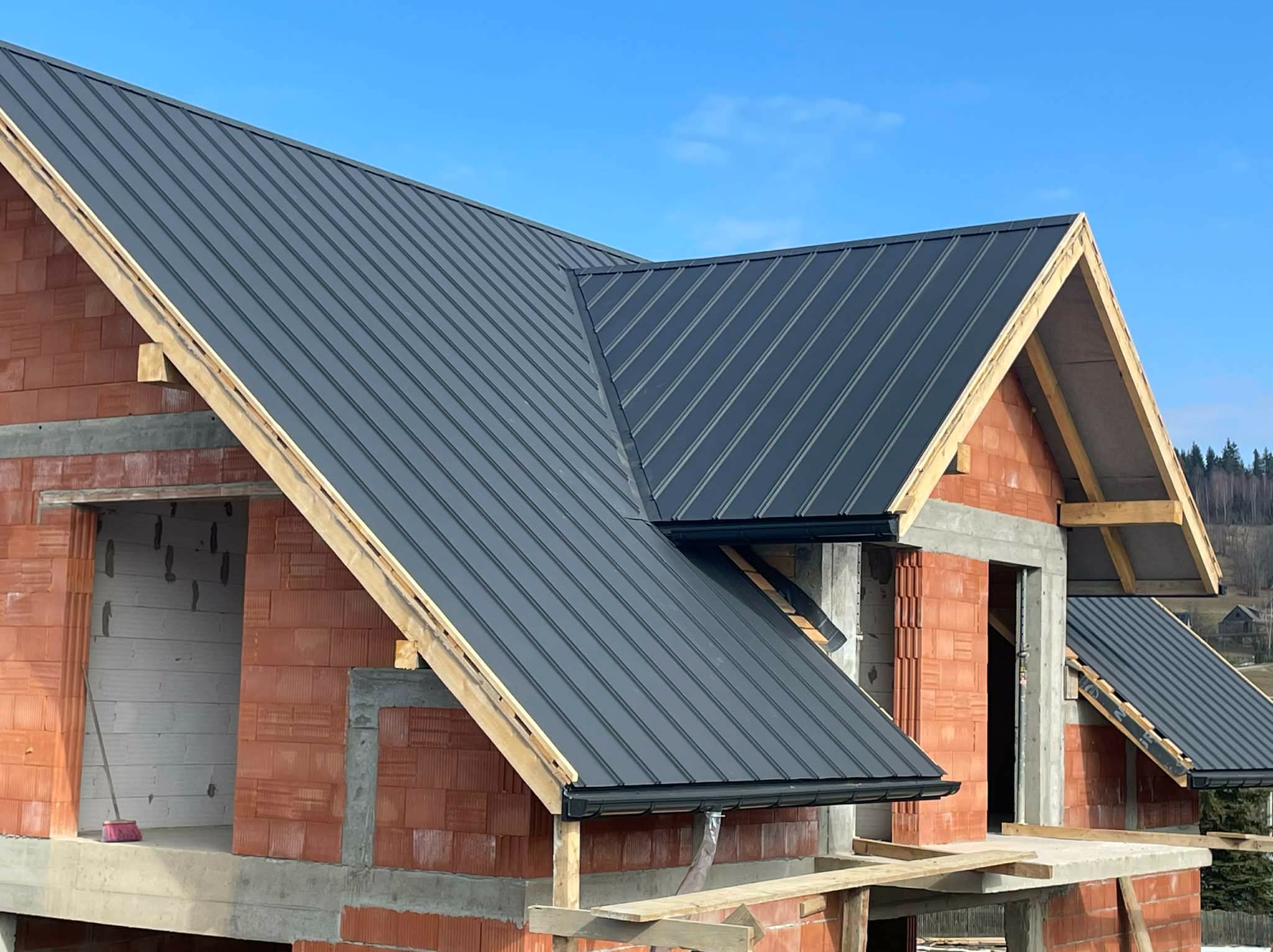 Nowy, grafitowy dach z blachy trapezowej na surowym budynku z czerwonej cegły. Widoczne rynny i drewniana konstrukcja dachu. Błękitne niebo w tle.