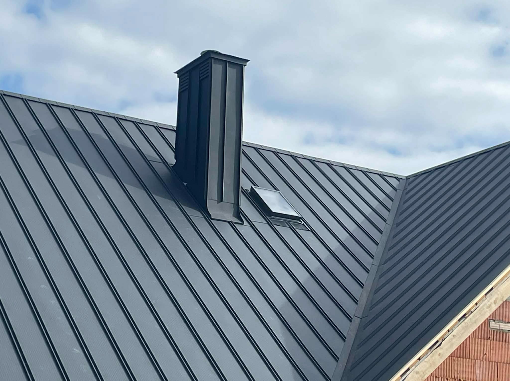 Grafitowy dach z blachy na rąbek stojący, z kominem i oknem dachowym, na tle pochmurnego nieba. Widoczny fragment muru budynku w surowym stanie.