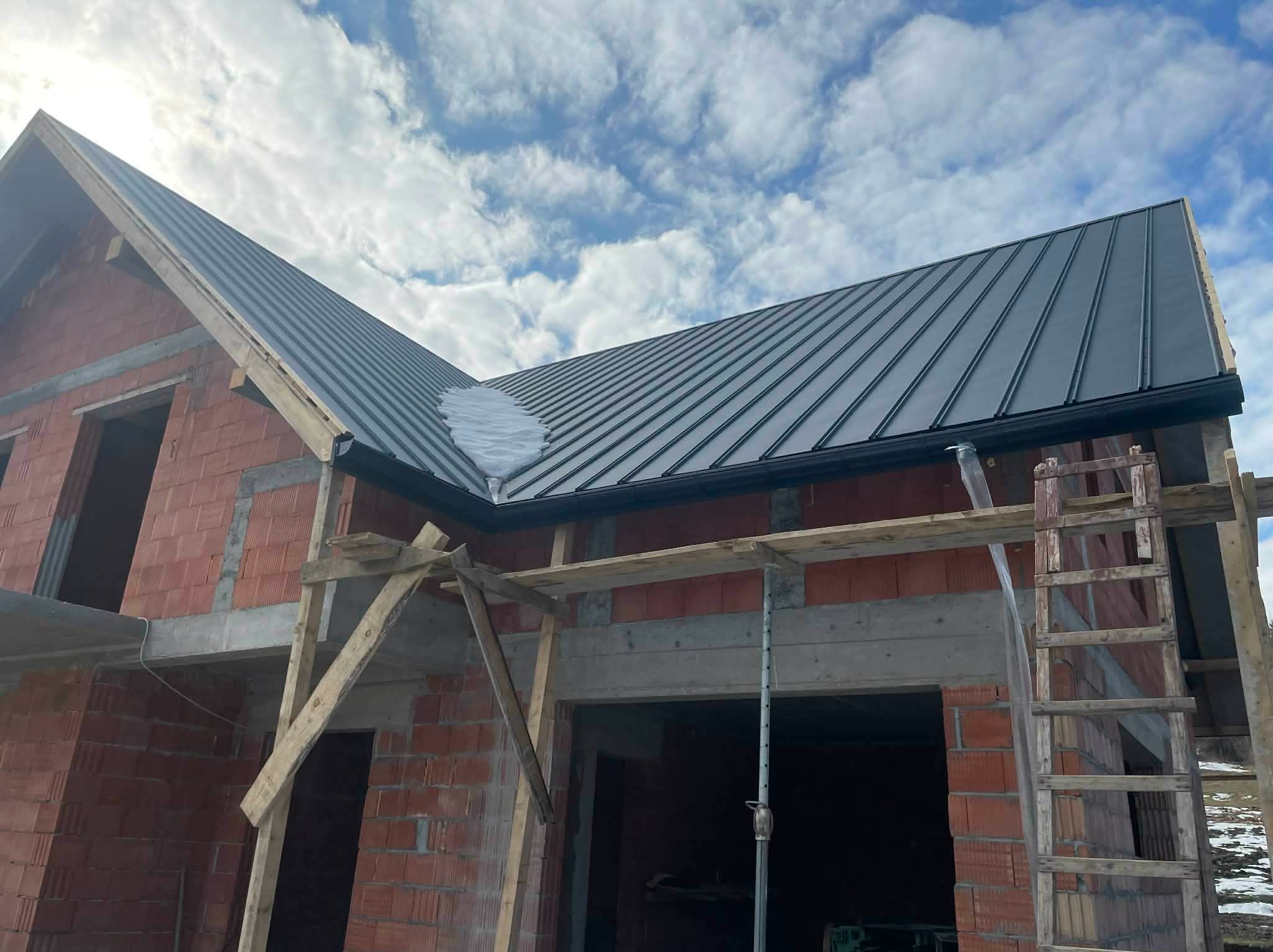 Nowy, grafitowy dach z blachy na surowym budynku w trakcie budowy, z widocznymi rusztowaniami i fragmentem śniegu na dachu. Błękitne niebo z chmurami w tle.