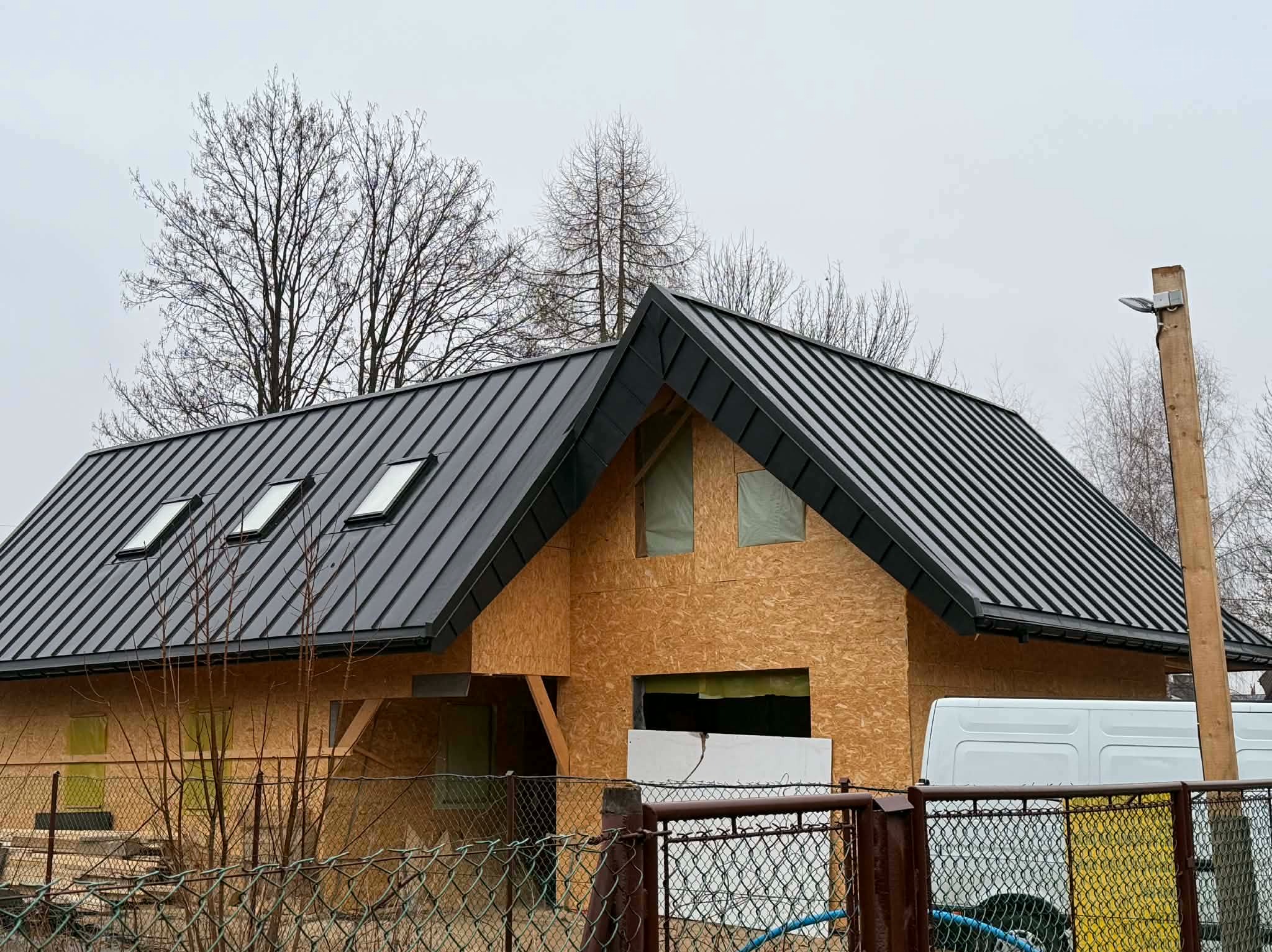 Dom w trakcie budowy z czarnym, metalowym dachem z trzema oknami dachowymi. Ściany z płyt OSB, widoczne elementy konstrukcyjne. Szara, pochmurna pogoda.