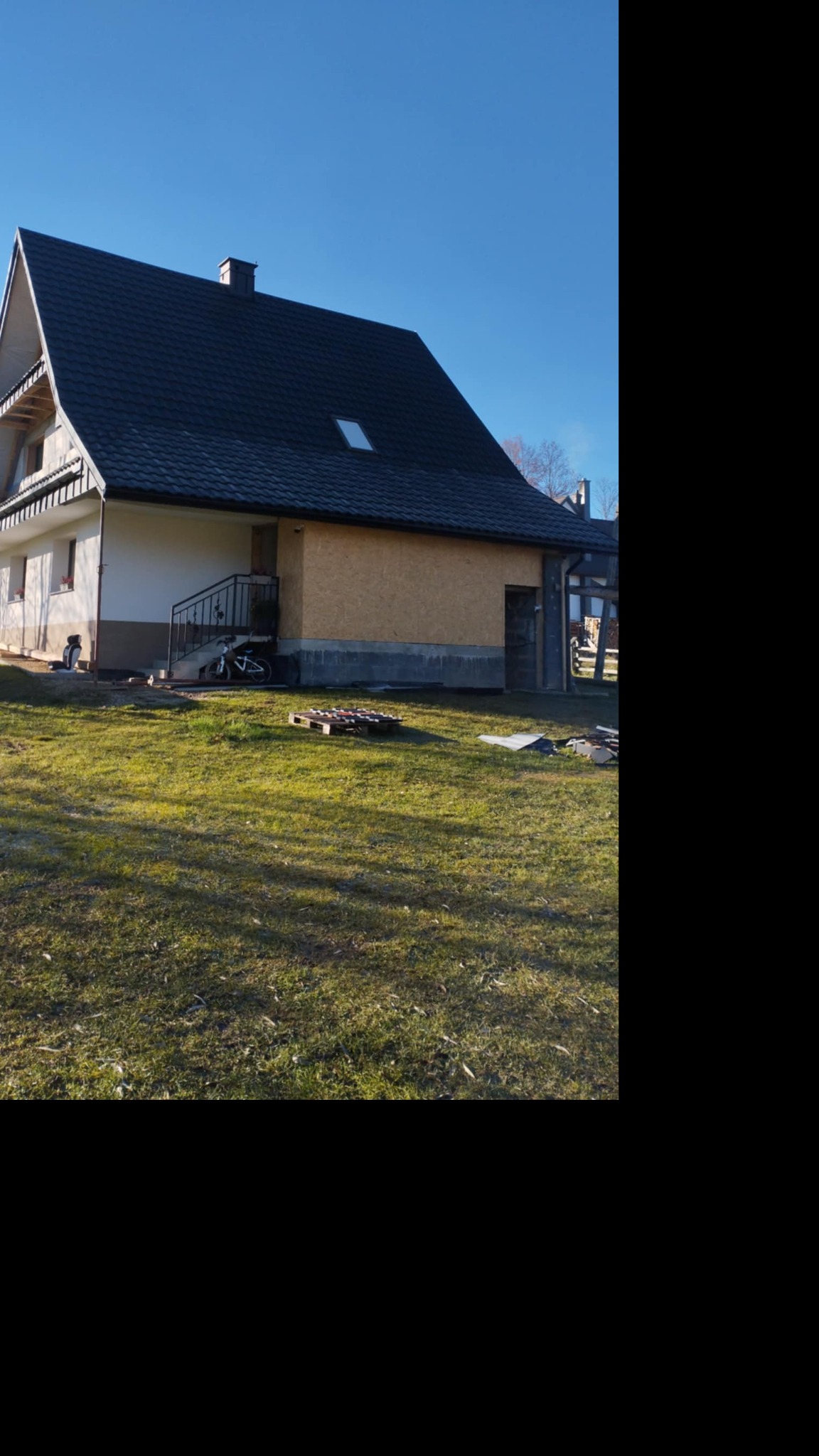 Dom z ciemnym, nowym dachem i oknem dachowym, widoczny z trawnika w słoneczny dzień. Część elewacji w trakcie wykończenia, widoczne palety i materiały budowlane.