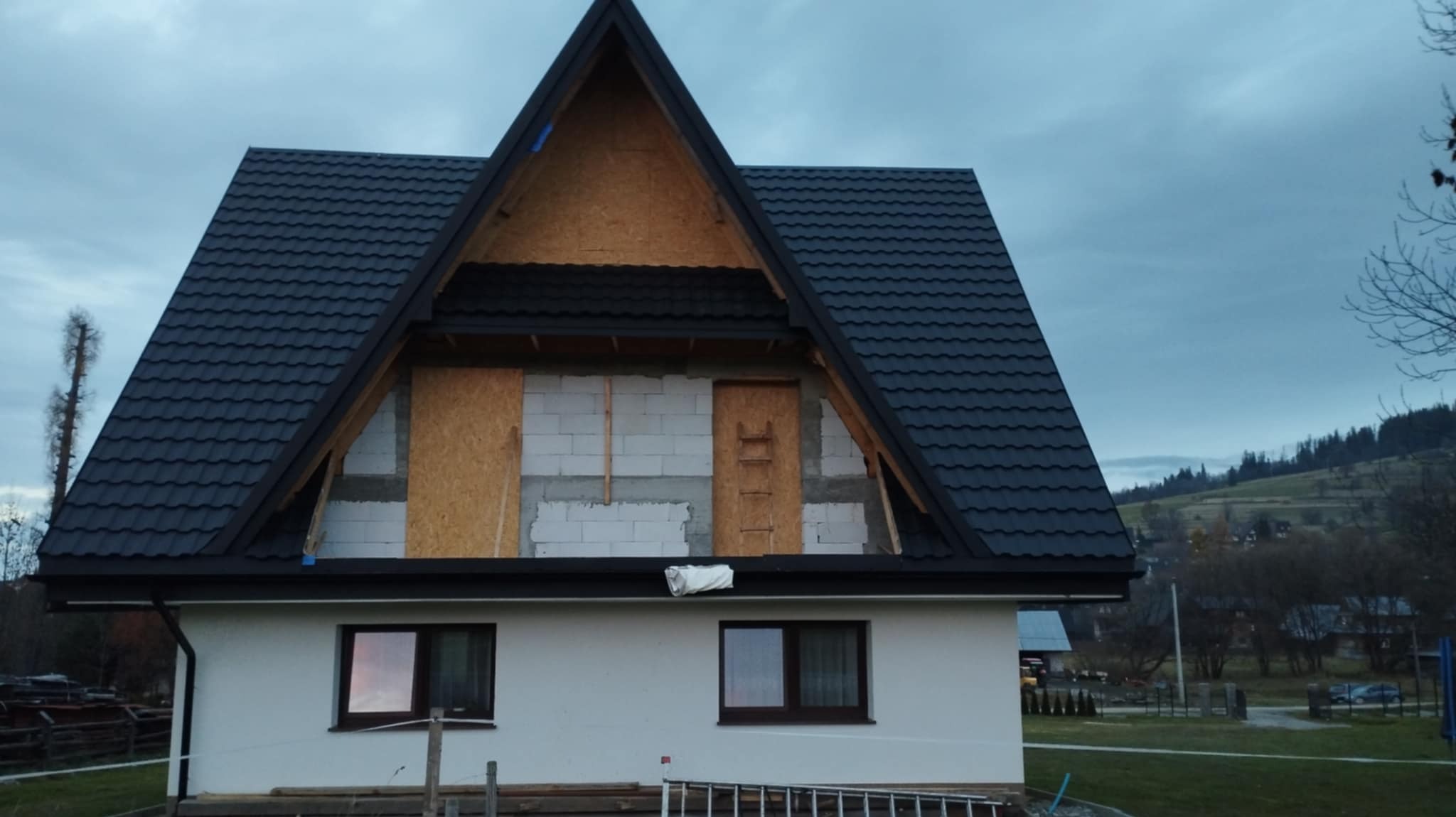 Dom w trakcie prac dekarskich: czarna dachówka kontrastuje z jasną elewacją. Widoczne elementy konstrukcyjne dachu i rusztowanie. Tło stanowią zalesione wzgórza.