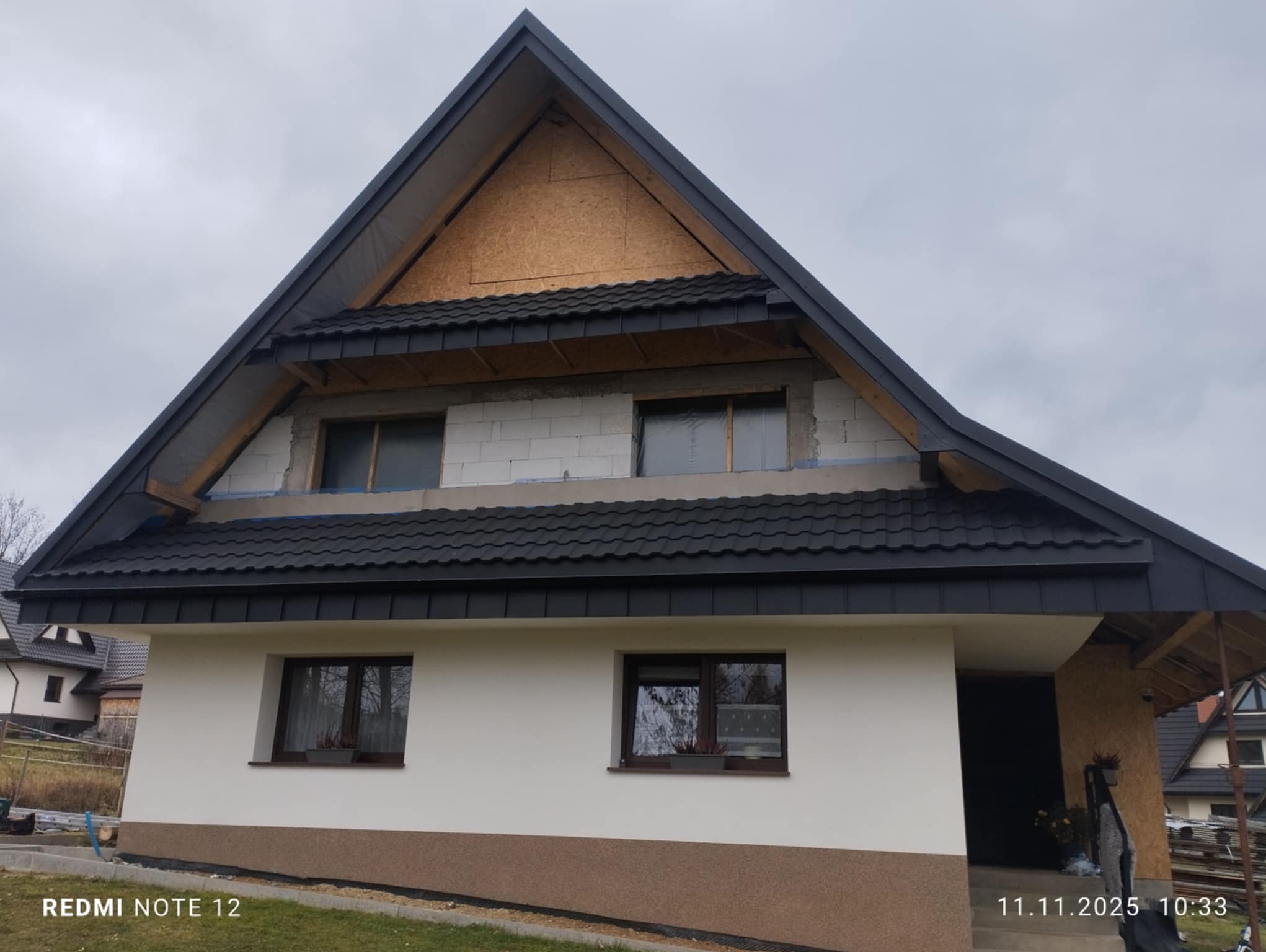 Dom w trakcie krycia dachu czarną dachówką. Widoczne fragmenty konstrukcji i ocieplenia. Częściowo otynkowana elewacja. Ujęcie z dołu, pochmurne niebo.