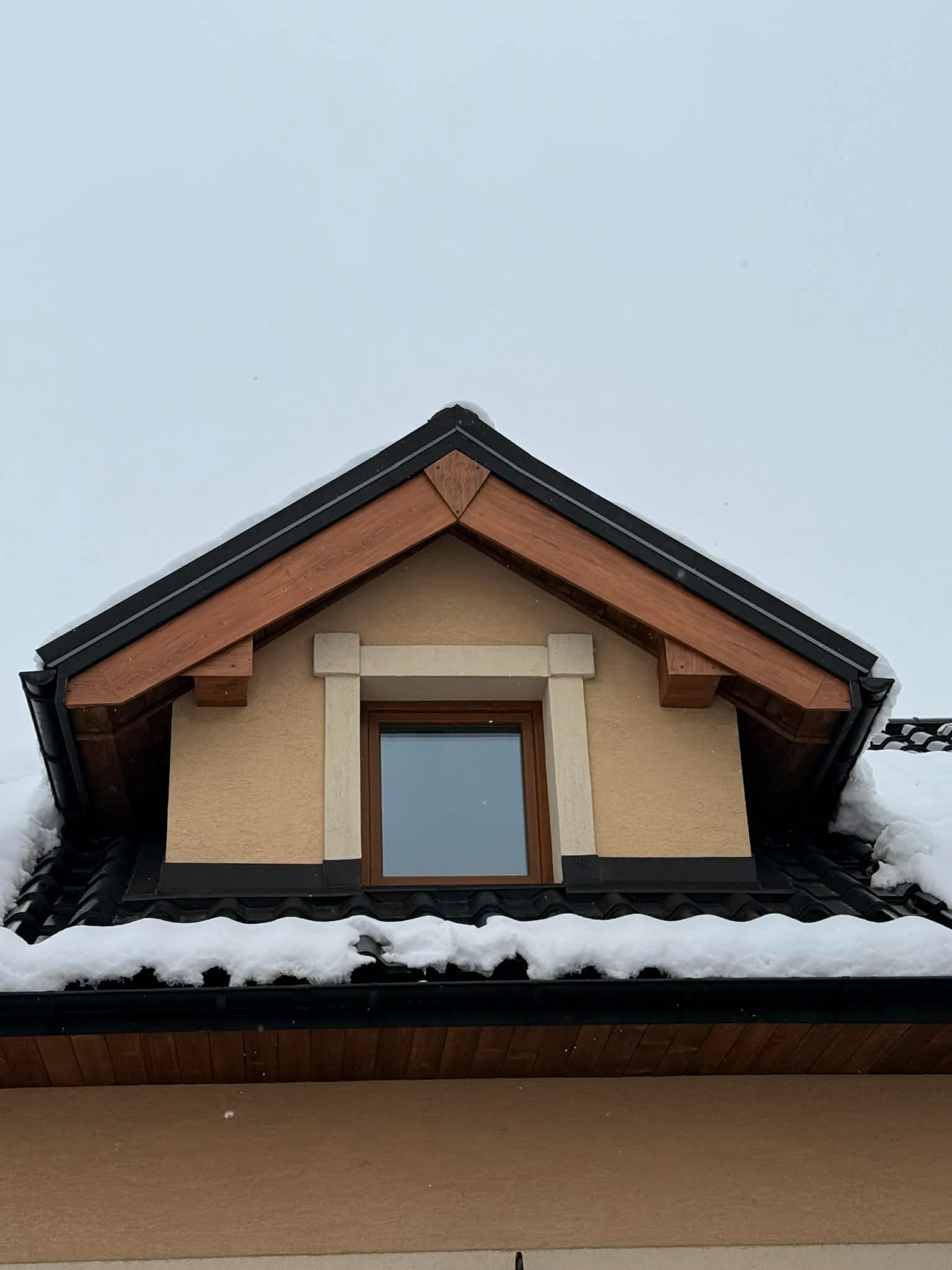 Zimowy widok na lukarnę z czarną dachówką i drewnianymi elementami wykończeniowymi. Śnieg na dachu i rynnach. Jasna elewacja budynku.
