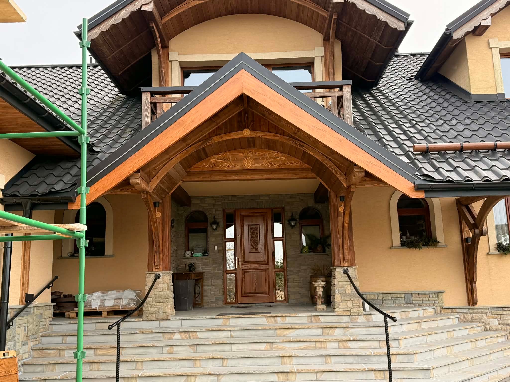 Dom z ciemną dachówką i drewnianym podcieniem nad wejściem, widoczne rusztowanie. Detale wykończenia z kamienia i drewna. Schody wejściowe z jasnych płytek.