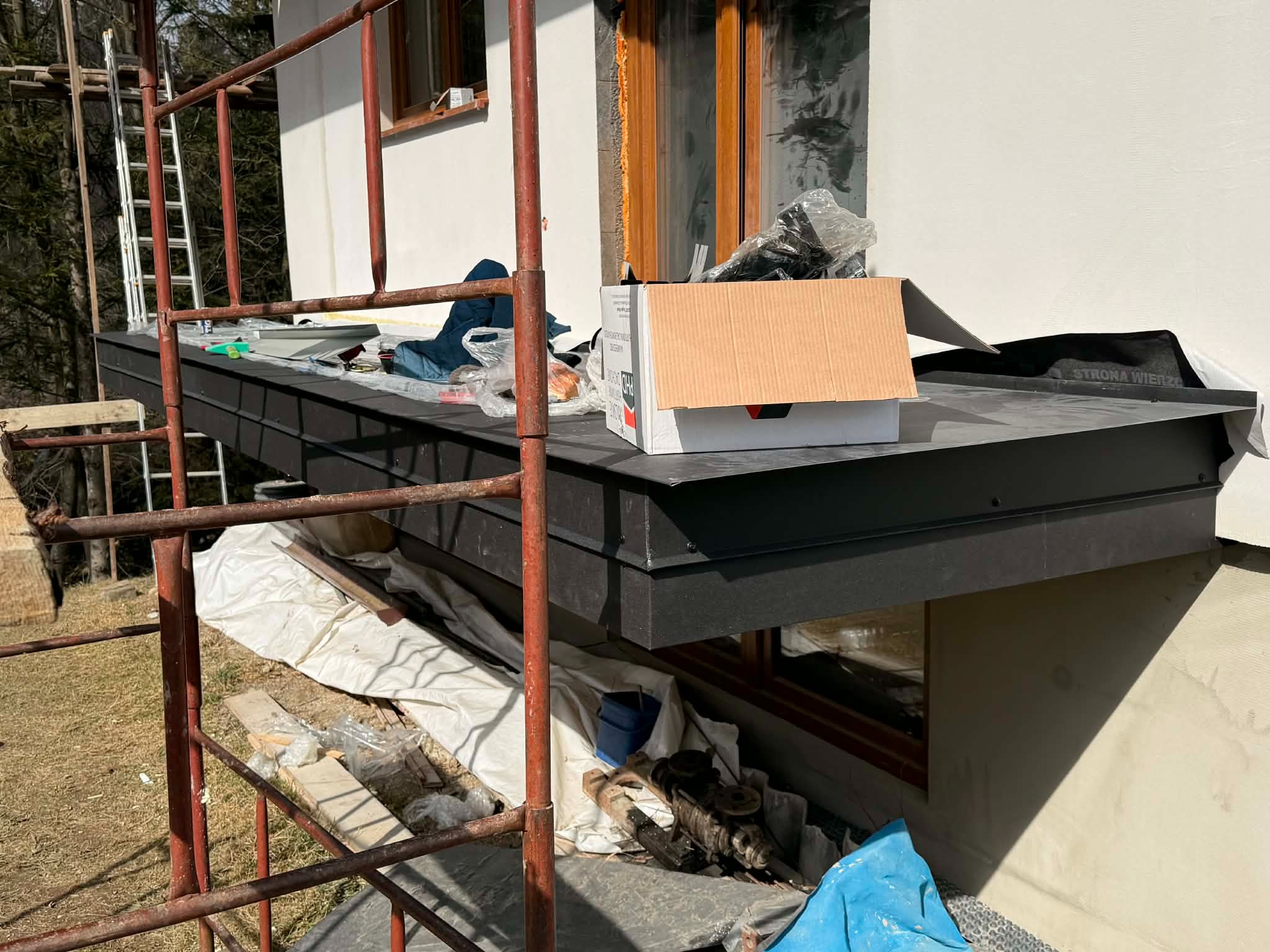 Czarny, płaski dach nad oknem piwnicznym w trakcie prac wykończeniowych. Widoczne rusztowanie, materiały budowlane i narzędzia. Dach pokryty czarną membraną.